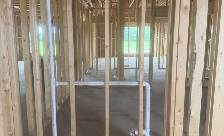 Upward view looking into open ceiling joists and roof trusses showing a long horizontal PVC plumbing pipe cleanly sloped and supported through structural timbers.