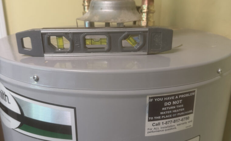 A gray A.O. Smith gas water heater tank with a small torpedo level resting flat on its top surface, verifying the alignment of the copper plumbing line connections and the galvanized exhaust flue above