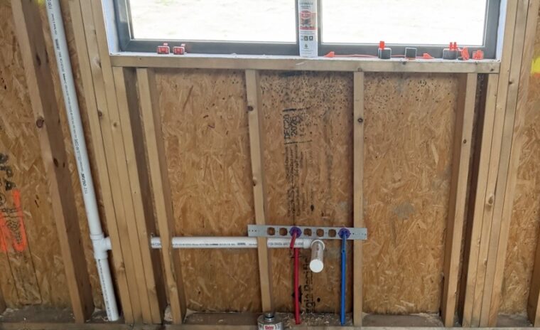 Framed wall with drain arm, PEX water lines, and a handwritten note on wooden stud framing.