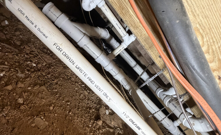 Tight crawl space view showing multiple PVC drain branches secured to joists with metal perforated strapping.