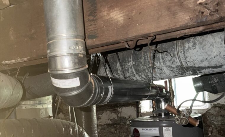 Double-wall B-vent flue line elbow connection routed from a Rheem gas water heater in the basement up through floor joists.