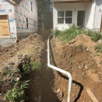 24AA2636 11A4 4C33 BC5F DC571952D7E9 b9cac80e 0e00 4b53 8c79 4170afd0eacf o Image 2 (Foundation connection): A close-up view of a white PVC sewer cleanout stack exiting a concrete home foundation and dropping down into an open dirt trench