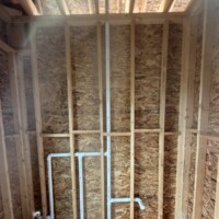 2F7E393A 6DED 4CC1 BC53 9B6146086E0D d4a2e8ab bfc6 4eaf ab21 400afee7541c : Interior wood framing of a house under construction showing a vertical PVC drain waste vent stack running through the floorboards and splitting into horizontal vanity branch lines.