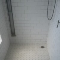 59DBB000 3355 4257 AFD4 ABB1BA1F09CE 14a68da6 d4cd 499b 8bb2 bc5c712c679a View of a white tiled shower floor with a circular stainless steel drain grid and chrome valve handles mounted on a subway tile wall.