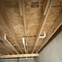 731B809C 261A 4C79 B42B 291DF5CE4624 c3be502f 8bfa 493b 8603 62369e77370e Unfinished basement joists with sloped main PVC drain run, waste lines, and structural supports.