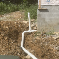 9005E01F 3CC2 4209 B02E 8381E9B1A22E cccc62d4 7a1a 4cef 9a3e bf7d854f556b o Image 4 (Riser junction): The termination point of the white PVC sewer line connecting to a vertical green main utility riser pipe next to an electrical transformer box