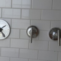 B2211610 7DA0 4CF5 A327 CF5F103B4862 33dc1061 6a81 4ccd 9f7f e9e3fe92f5ed Close-up view of three modern, cylindrical chrome shower control valve handles mounted horizontally on a white subway-tiled wall.