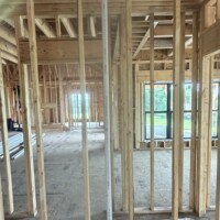 CB17E9E9 3851 4B28 B391 1B835DEAC7EB b03b4c68 570b 4a77 8836 b520f3fb2a17 Looking through a framed partition wall showing a vertical PVC utility pipe running down to a clean hole cut in the subfloor, with green fields visible out the large windows