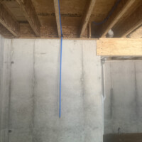 ReactNativeBlobUtilTmp 1vyqmuzyf4syqoapykc7t Concrete basement foundation wall with vertical PVC drain stack and overhead red and blue PEX piping.