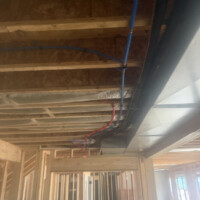 ReactNativeBlobUtilTmp 7zkgq3ui3aul3w7sml88 Overhead red and blue PEX water supply lines clipped securely to ceiling joists in framing. • Image 4: Shower rough-in framing showing red and blue PEX lines connected to a brass mixing valve with PVC vent.