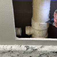 ReactNativeBlobUtilTmp j4hqzrfgfc79brfxsghy9p Drywall cutout exposing damaged PVC drain stack line and separated branch tee behind laundry room wall.
