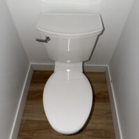 ReactNativeBlobUtilTmp 3574vbughut7spg5f88ca White two-piece elongated toilet installed on wood-look flooring with chrome flush lever.