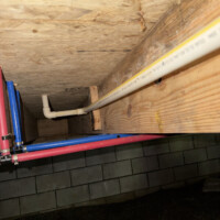 ReactNativeBlobUtilTmp 3mvehwfg3c4b77zrk6xwe6 Open crawlspace framing view showing white PVC DWV drain piping with cleanout plug running alongside PEX lines.