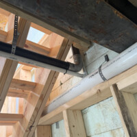 ReactNativeBlobUtilTmp ed6s4y1vgwq4dn07nil1l5 Looking up through open floor joists showing black iron gas lines, PEX water lines, and PVC drain pipes strapped to framing.