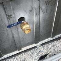ReactNativeBlobUtilTmp ijeux3m8xvjzljg8f63lp Exterior ground-level shot of brass ball valve hose bib stubbed out through dark metal siding over gravel base.